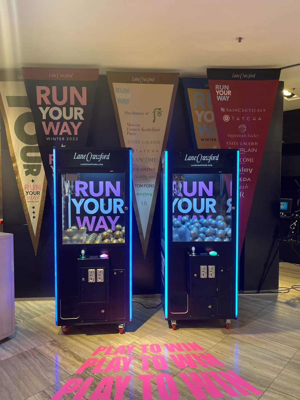 商場活動中一對黑色夾公仔機,機身與玻璃印有白色 RUN YOUR WAY 字樣,機身邊緣亮起藍色燈條,地上投射粉紅色字句,背景懸掛三角旗