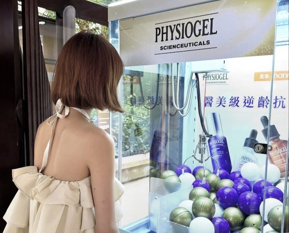一部透明機身的夾公仔機,頂牌印有護膚品牌標誌,機內背板印有產品圖片及中文宣傳字,機內放滿紫色、金色及白色膠球,一位女士正在按操作台的按鈕
