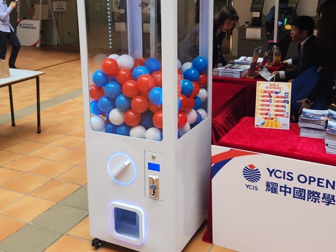 白色方扭蛋機放在學校開放日的攤位旁，機內裝滿紅、藍、白三色扭蛋，旁邊桌上放著換領告示牌