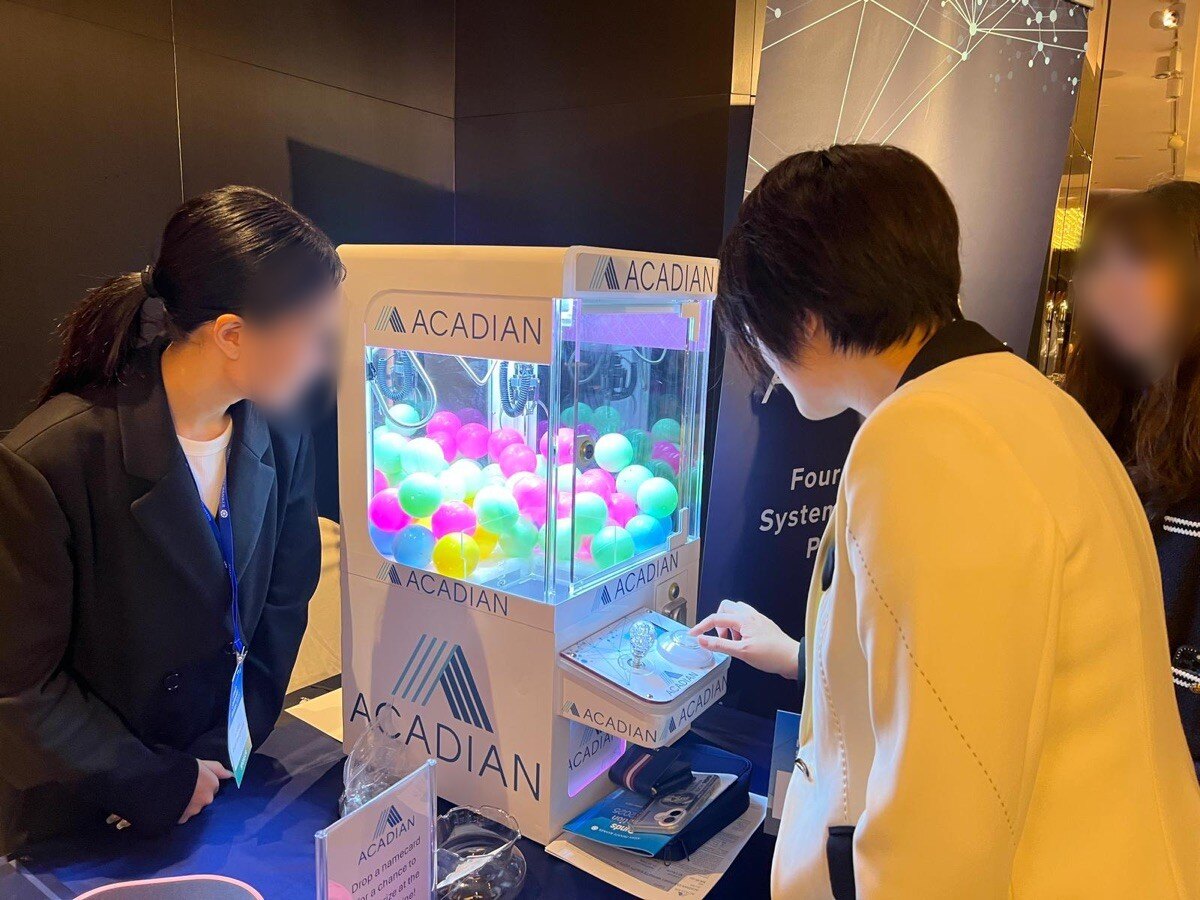展覽攤位的枱面上放著一部白色機身的桌上夾公仔機,機身印有公司標誌,機內放滿粉紅、綠、黃色膠球,一位參觀者正在按搖桿,攤位職員在旁觀看,枱上有收集名片的膠座